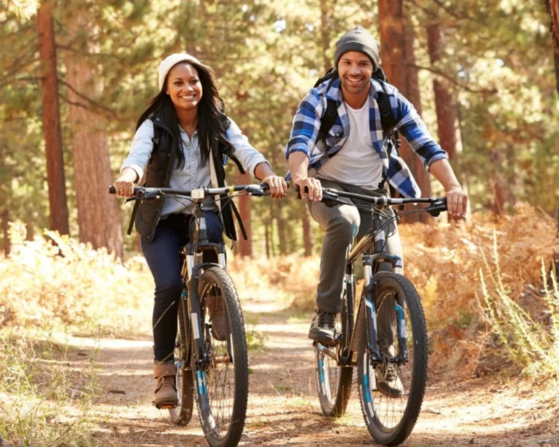 A smiling couple riding mountain bikes side-by-side along a sunny forest trail