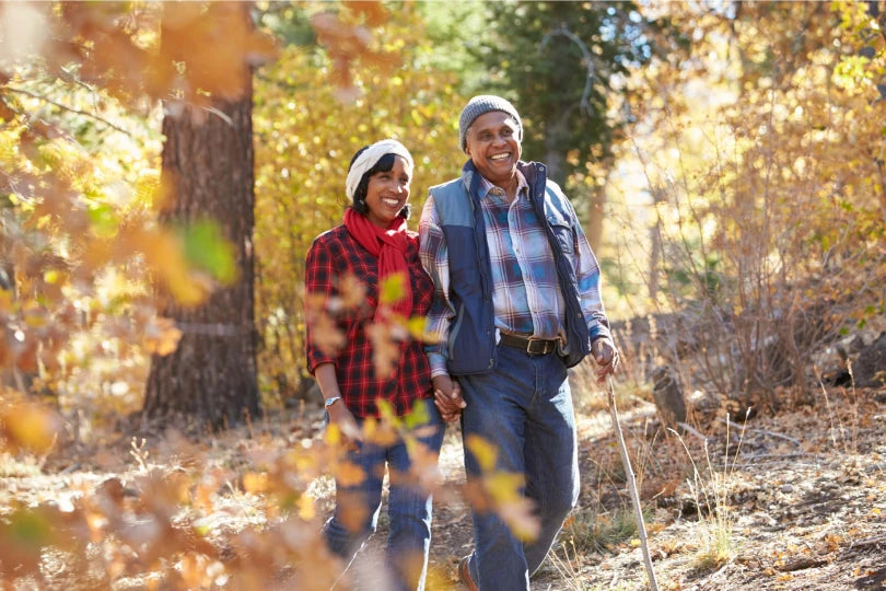 An older Black couple smiling and holding hands while hiking along a sunlit autumn forest trail